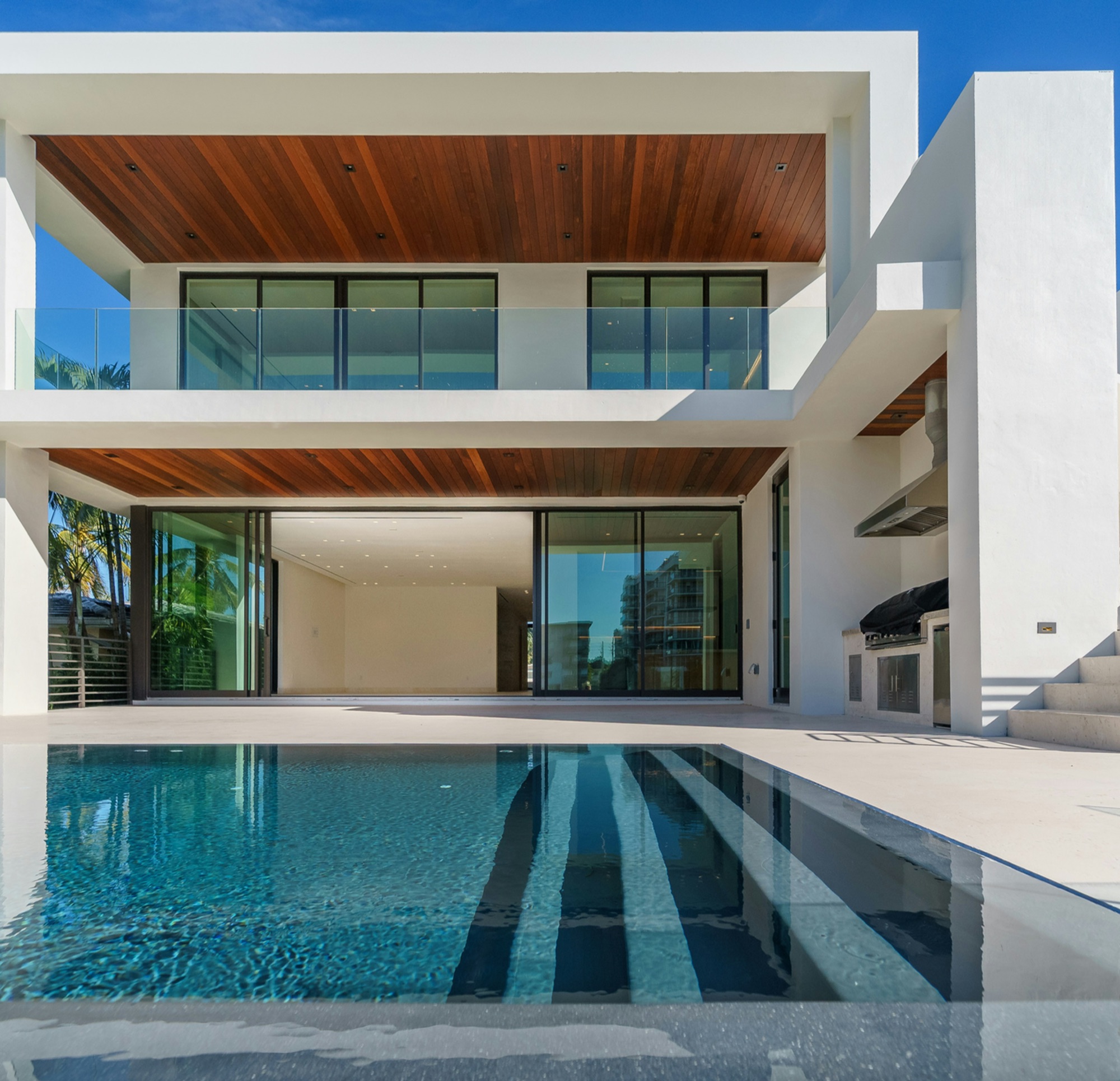 Modern white luxury home exterior with wood ceilings, large windows, and a clear blue swimming — Vireo Homes